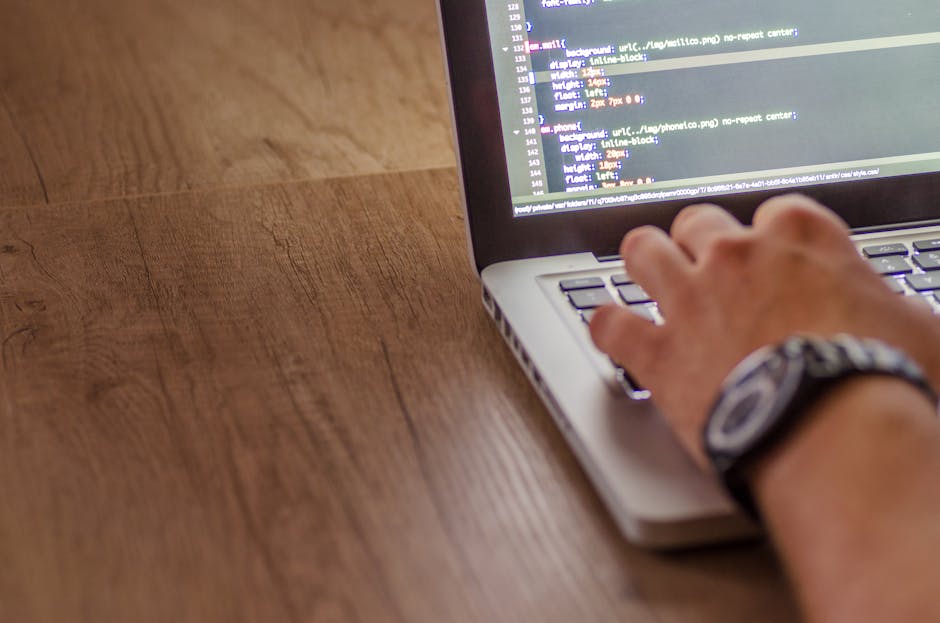 A person uses a laptop on a wooden surface, focusing on the screen displaying lines of code in various colors on a dark background. The individual’s right hand rests on the keyboard, with fingers po