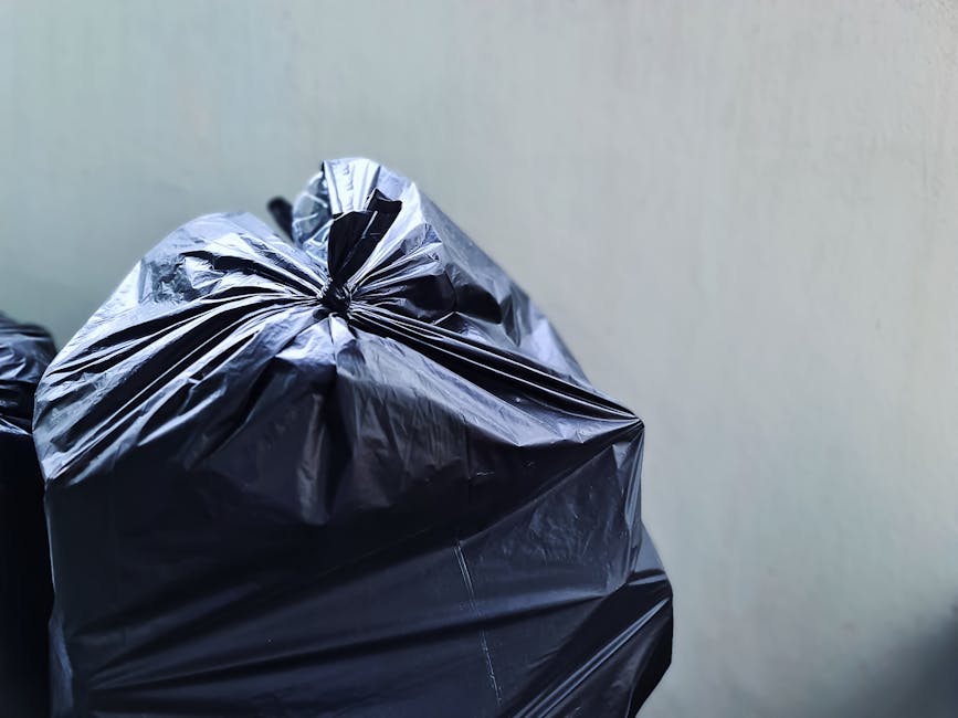 A black metal public rubbish bin situated on a cobblestone pavement in an urban setting, overflowing with various waste items. The top of the bin is covered with discarded paper cups, a plastic cup wi